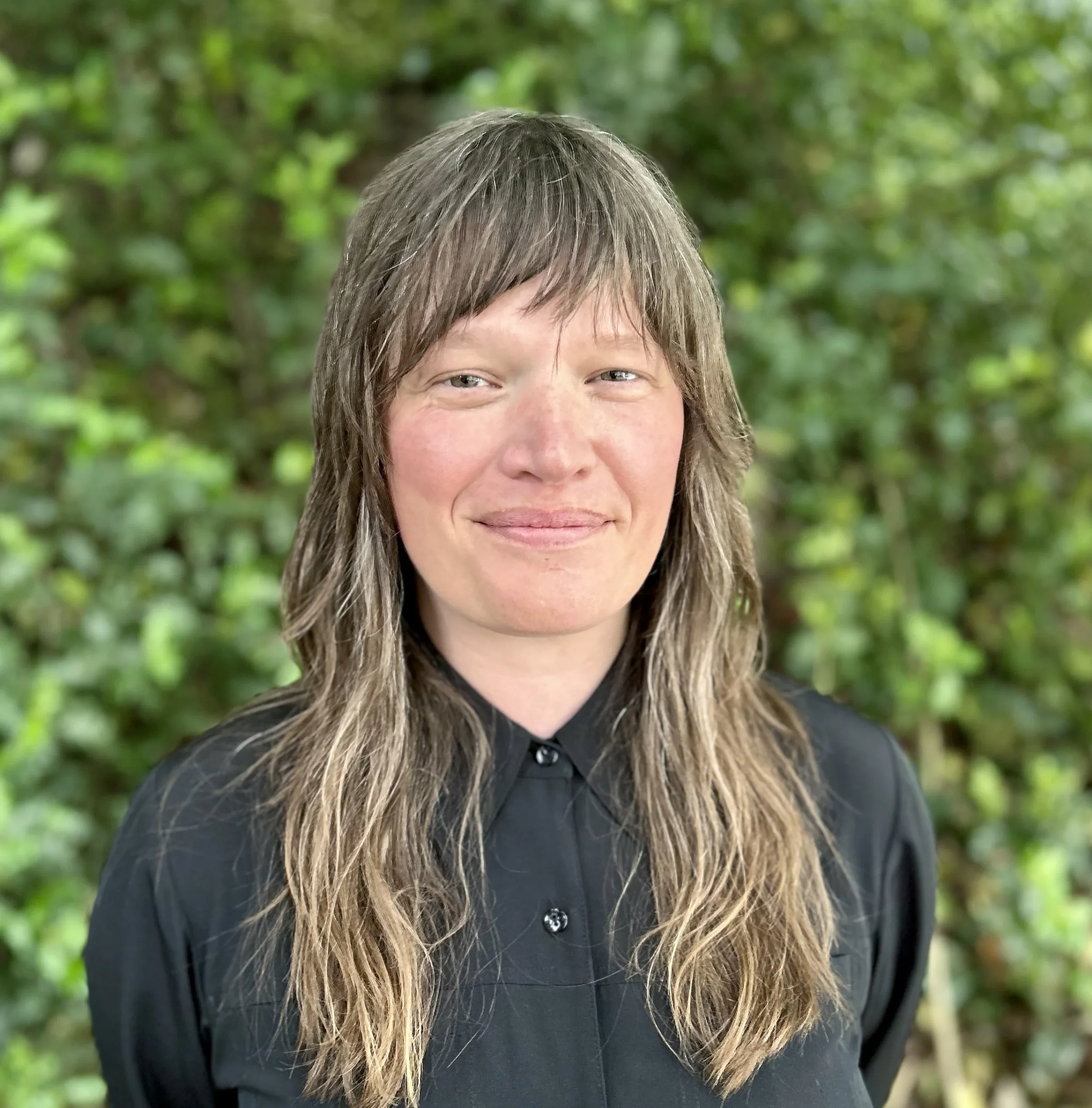 A woman with long, wavy brown hair and fair skin, wearing a black button-up shirt, standing outdoors with blurred green foliage in the background.