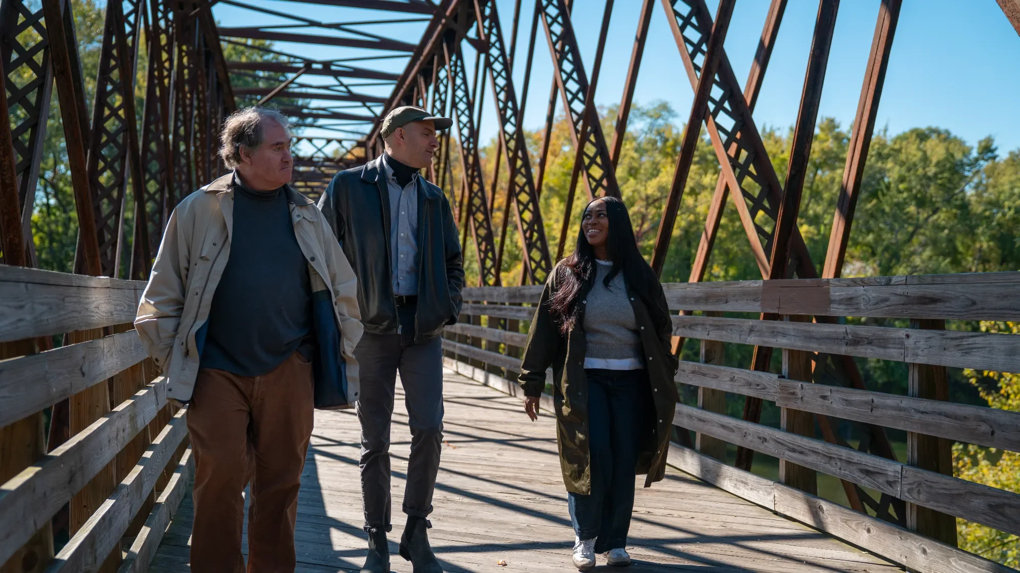 Three people walking on a wooden bridge with metal trusses, surrounded by green trees and a clear blue sky. They are talking and smiling, dressed in casual autumn clothing.