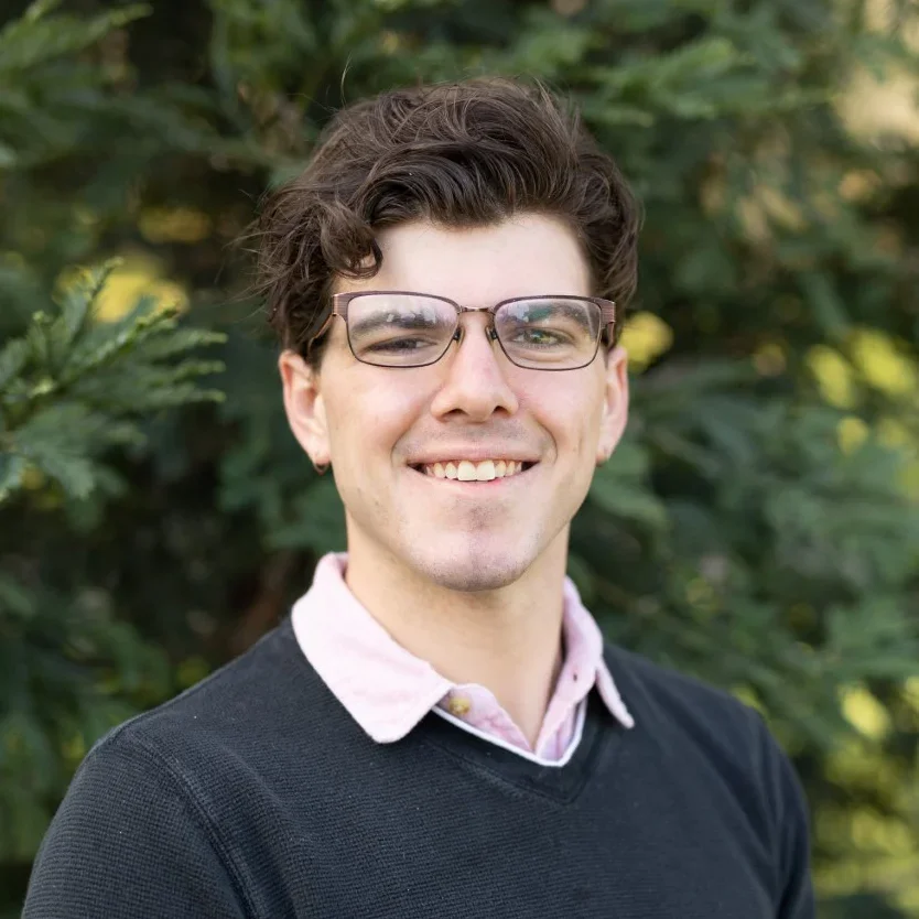 A young man with dark hair and glasses smiling outdoors, with greenery in the background.