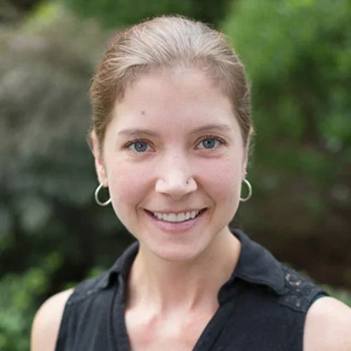 A woman with light skin, blue eyes, and brown hair tied back, smiling outdoors with green foliage in the background.