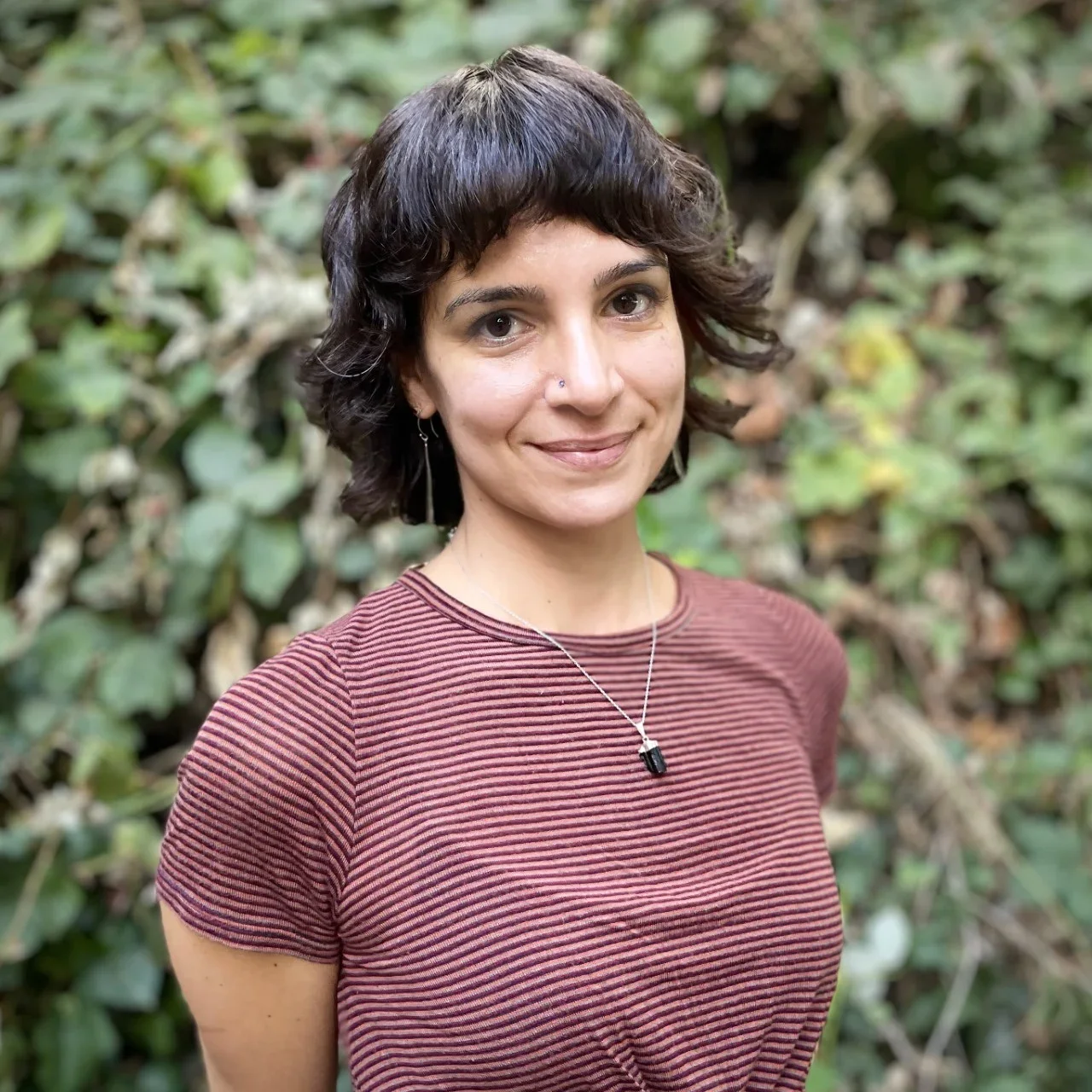 A woman with short, wavy dark hair and a nose piercing, wearing a striped maroon shirt, smiling outdoors with green foliage in the background.