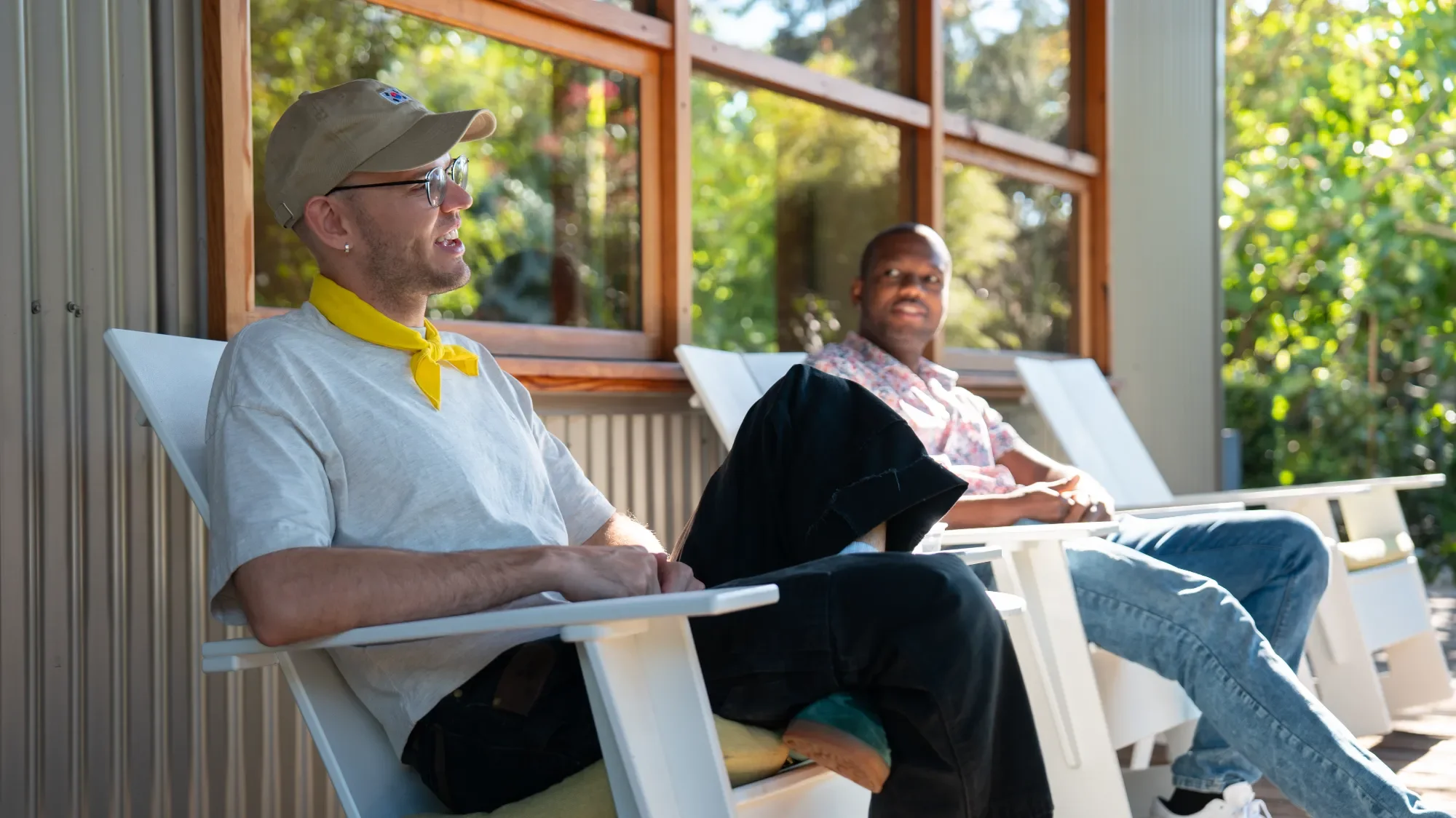 Two men sitting on white outdoor lounge chairs on a porch with large windows and greenery outside, engaging in conversation on a sunny day.
