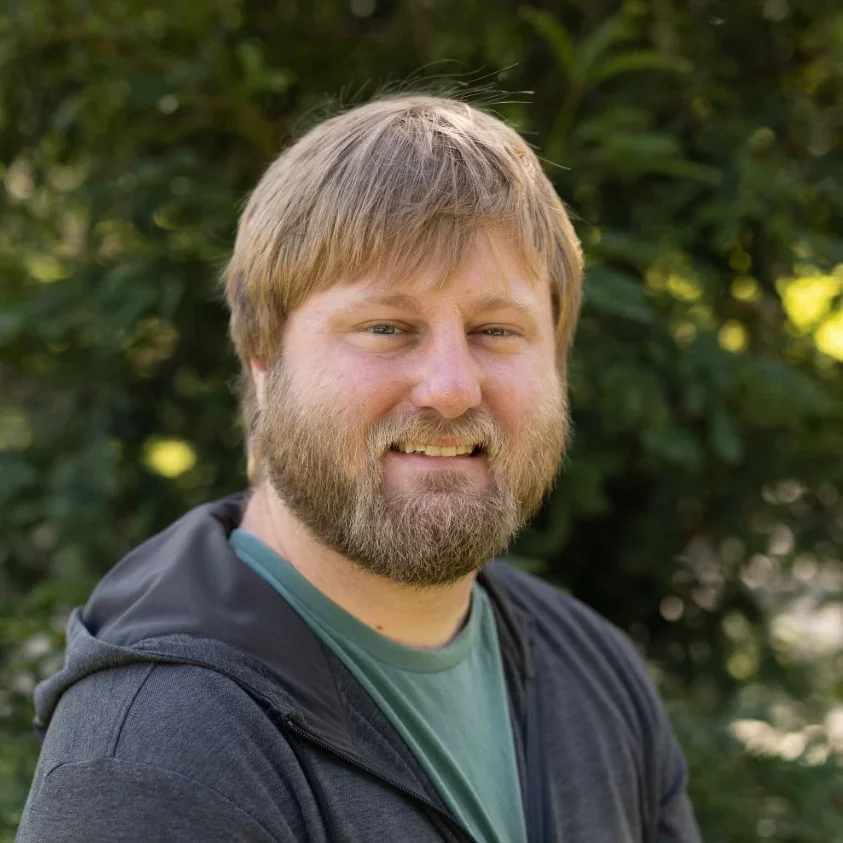 A man with a beard and mustache smiling outdoors, wearing a green t-shirt and a black zip-up jacket with trees in the background.
