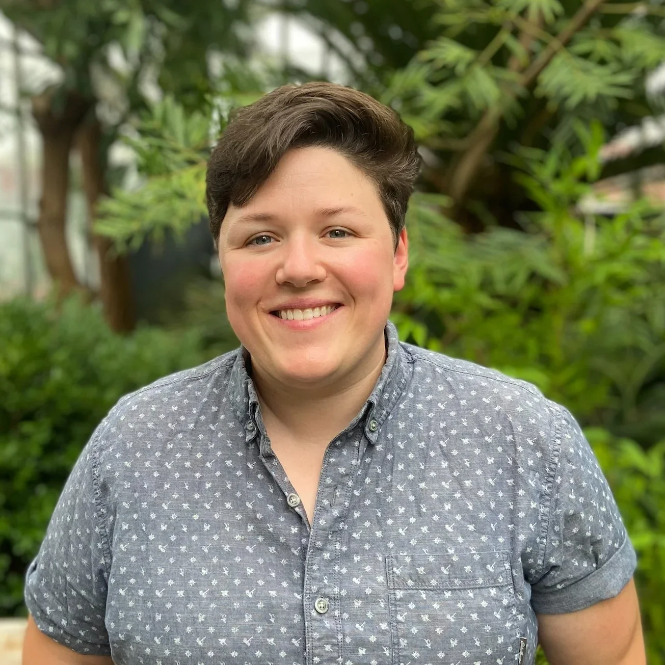 A person with short, dark hair smiling at the camera, wearing a gray patterned button-up shirt with rolled-up sleeves, standing outdoors with green foliage in the background.