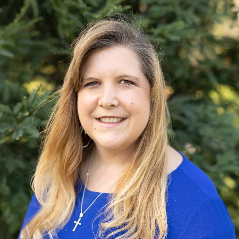 A woman with long blonde hair smiling outdoors with green foliage in the background, wearing a blue top and a cross necklace.