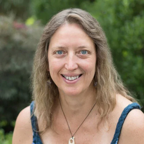A woman with long, wavy blonde hair and blue eyes smiling outdoors with green foliage in the background.