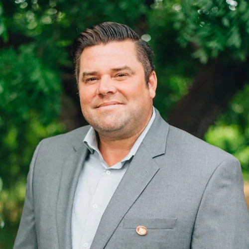 Portrait of a man in a grey suit outdoors with green trees in the background.