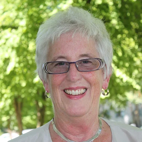 An elderly woman with short gray hair, glasses, and earrings, smiling outdoors with green trees in the background.