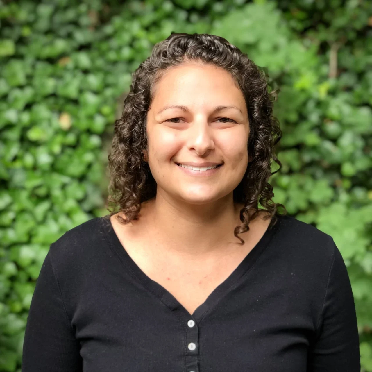 Portrait of a woman with curly hair, smiling, wearing a black button-up shirt, outdoors with green leafy background.