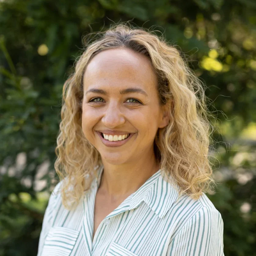A woman with curly blonde hair smiling outdoors against a background of green foliage, wearing a white shirt with thin light blue stripes.