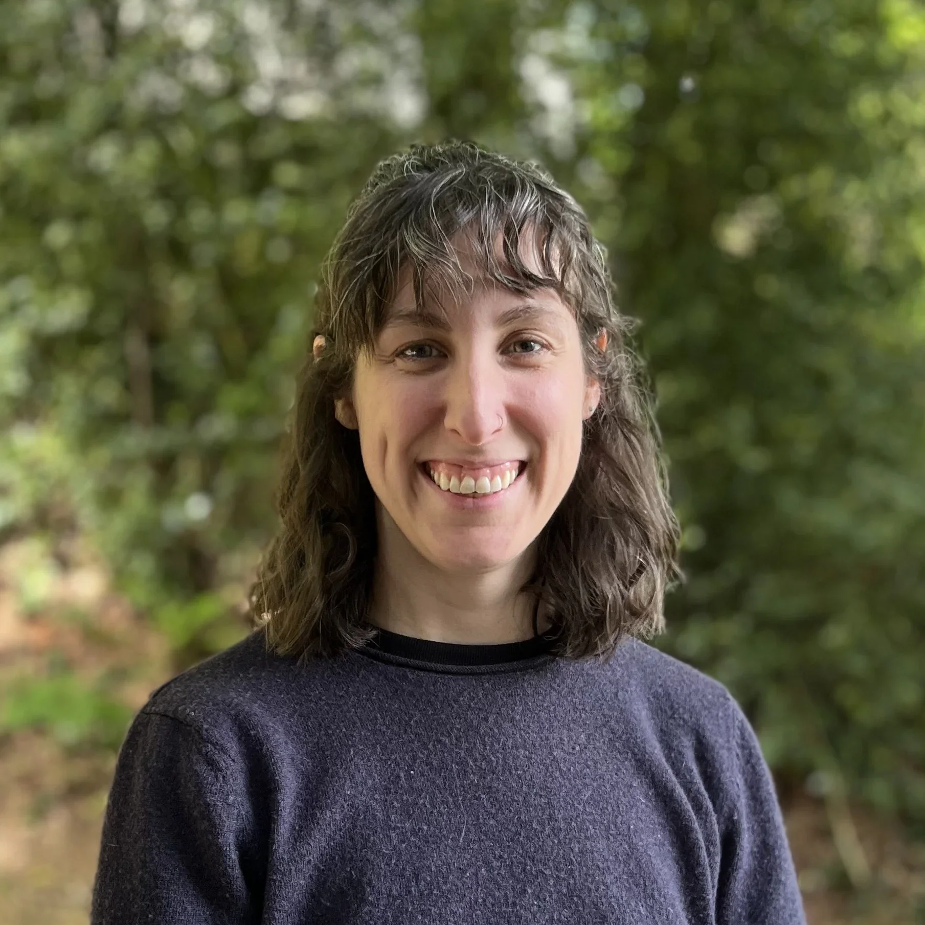 A woman with shoulder-length curly brown hair, smiling, wearing a dark sweater, standing outdoors in front of green trees.