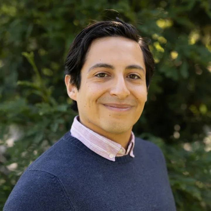 A young man with dark hair and light skin smiling outdoors, wearing a navy sweater over a light pink collared shirt, with green foliage in the background.