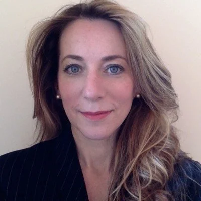 Portrait of a woman with long blonde hair, blue eyes, and fair skin, wearing a black striped blazer and pearl earrings, smiling gently at the camera.
