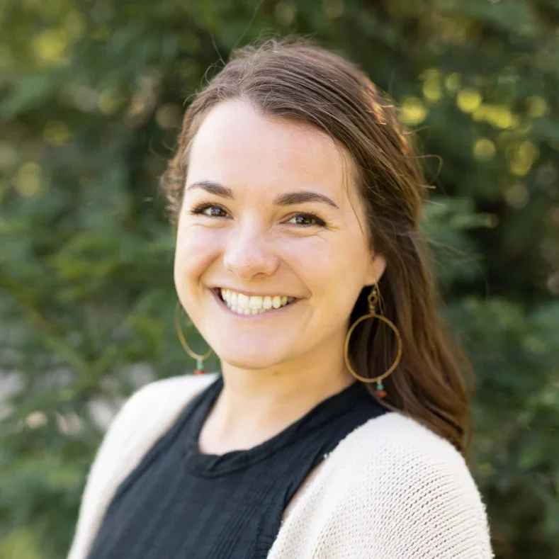 A young woman smiling outdoors with green trees in the background, wearing large hoop earrings and a black top with a white cardigan.