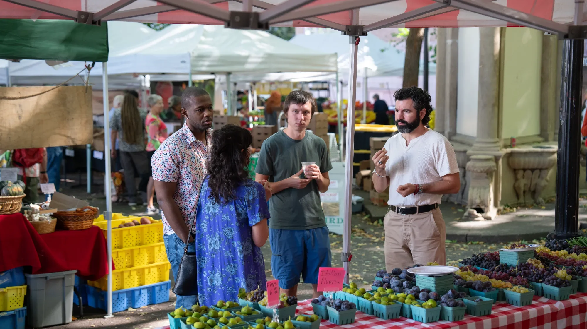 Four people are gathered at a produce stand selling figs and Italian plums at an outdoor market. The vendor is explaining the fruit to three customers, one man and two women, with a man holding a cup and gesturing.