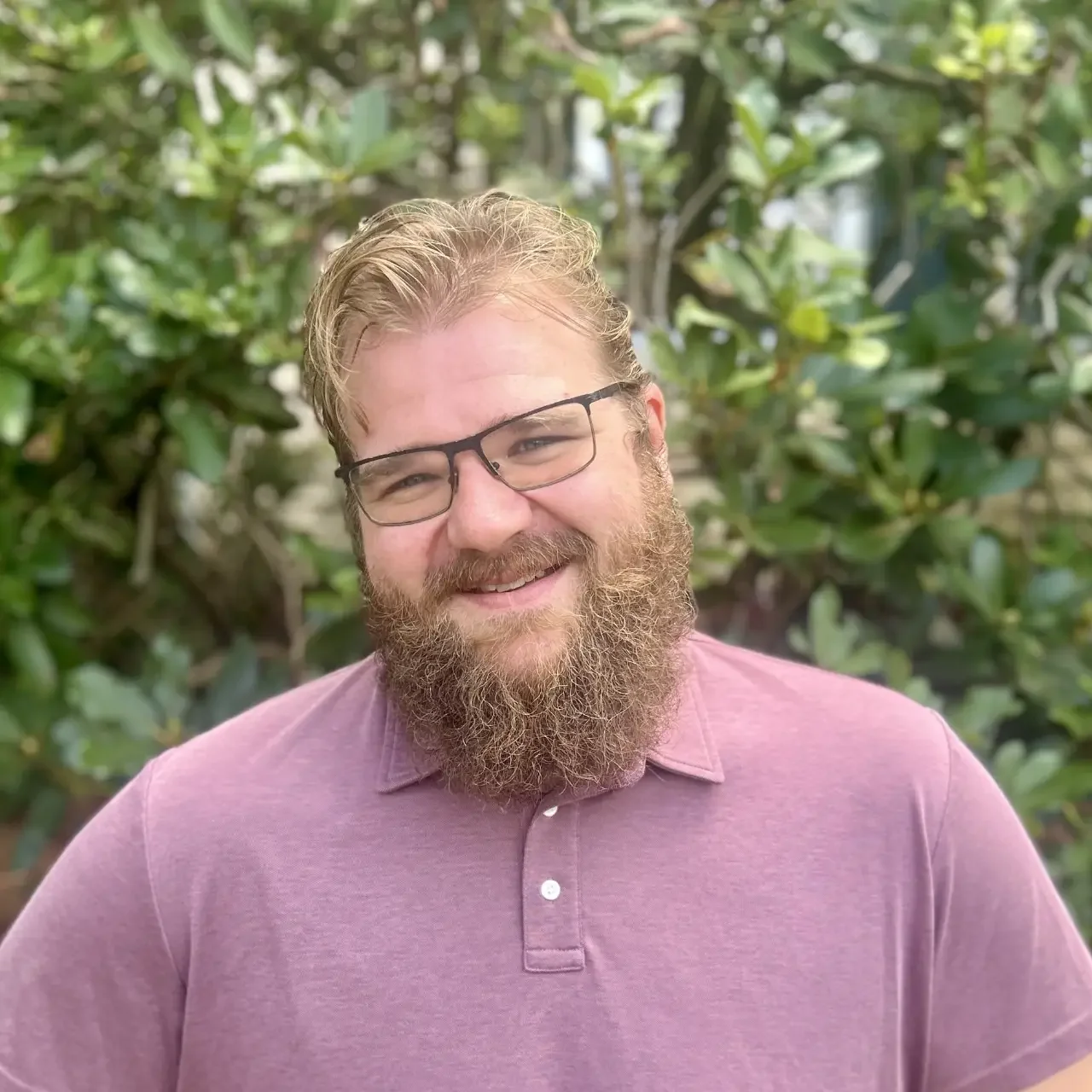 A man with glasses and a beard smiling outdoors, wearing a dark jacket over a hoodie, standing in front of green bushes.