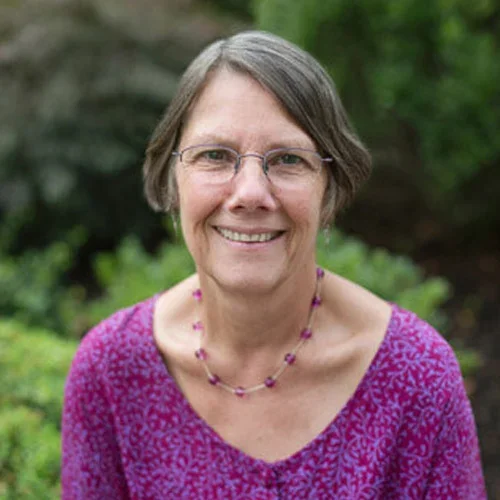 Smiling woman with short gray hair wearing glasses, a purple patterned top, and a purple beaded necklace, outdoors with green foliage in the background.