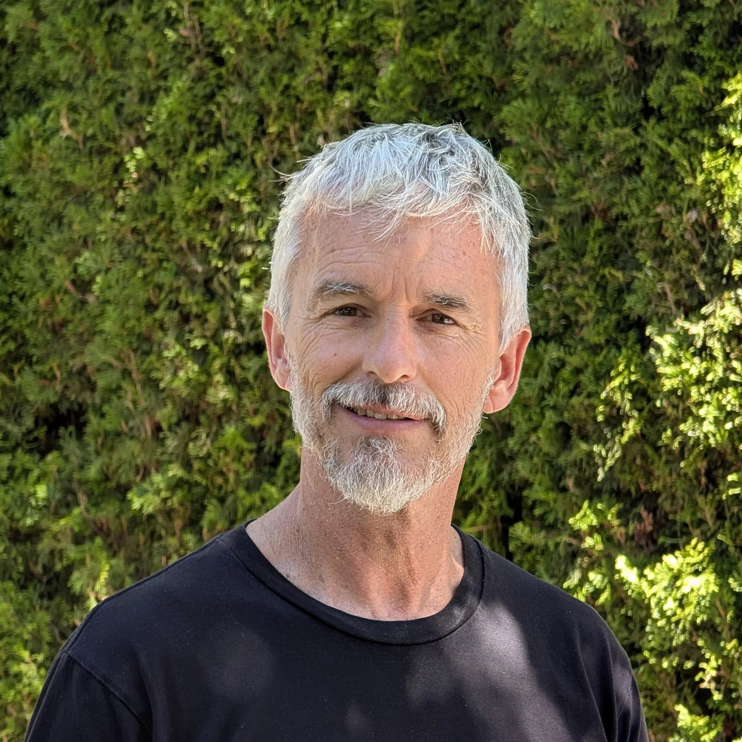 A middle-aged man with gray hair and beard, wearing a black shirt, standing outdoors in front of a green hedge.