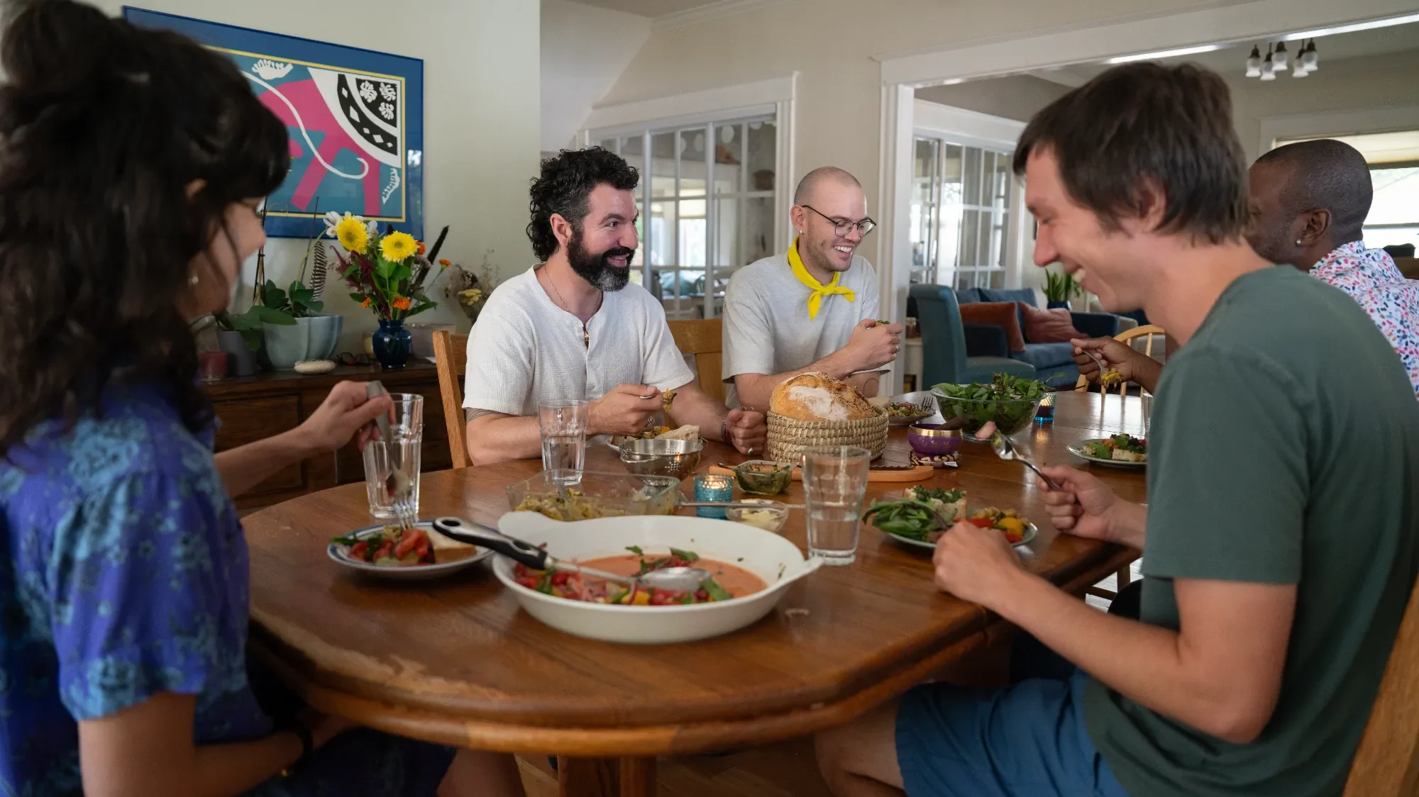 Five people gather around a dining table enjoying a meal, with food and drinks present, in a cozy, well-lit room.