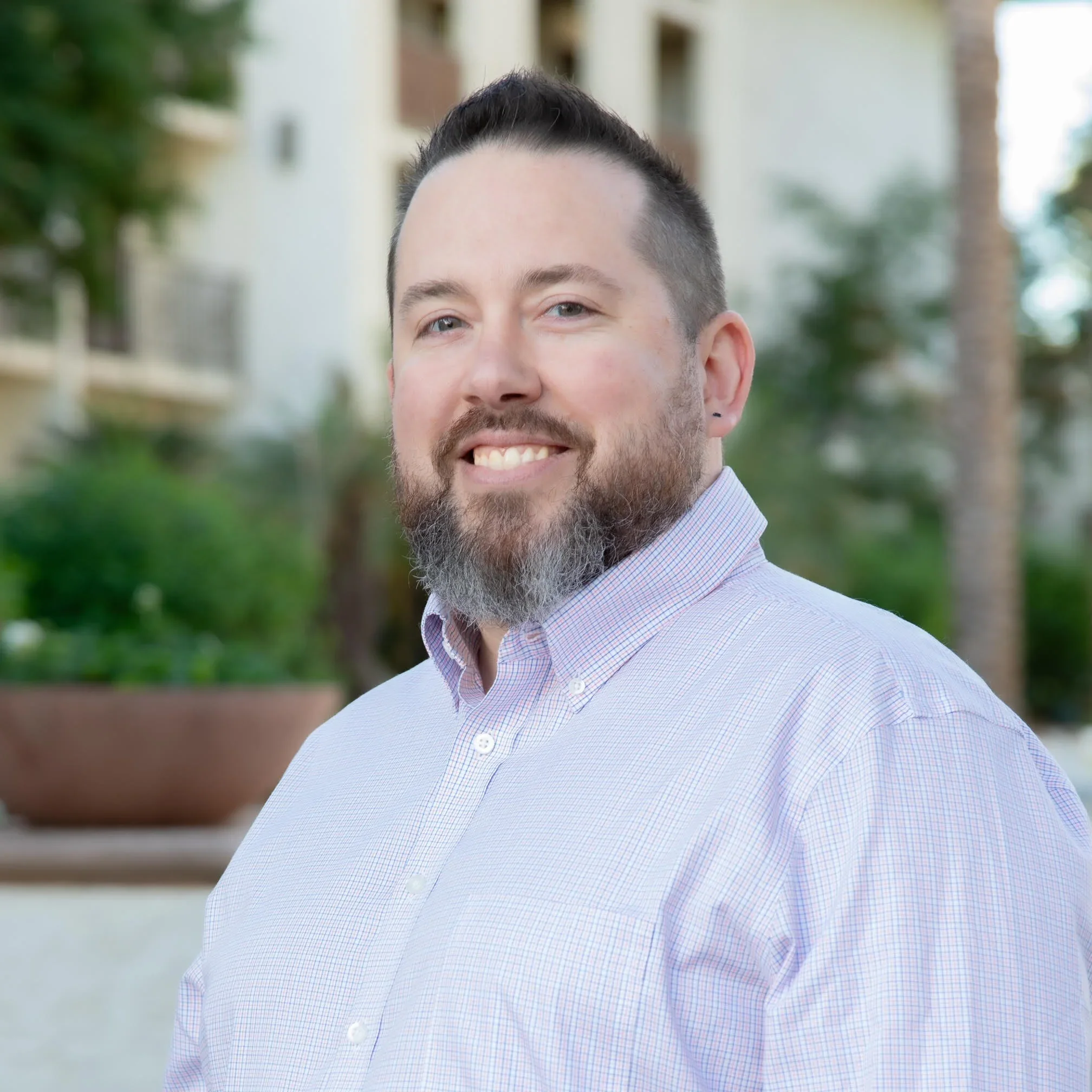 A smiling man with a beard and mustache wearing a light purple checkered dress shirt outdoors with blurred greenery and buildings in the background.