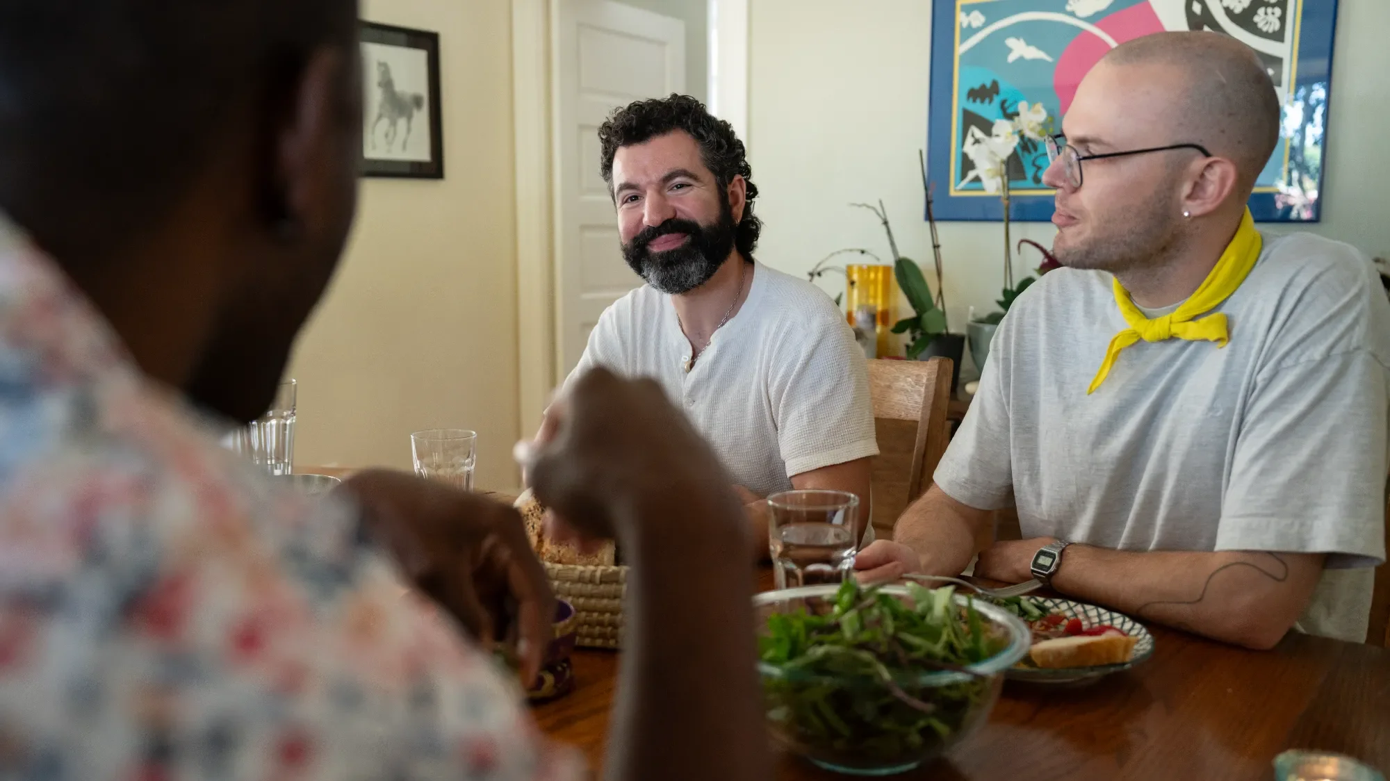 Three men sitting at a dining table with food and drinks, engaging in a conversation in a well-lit room with art on the walls and plants in the background.
