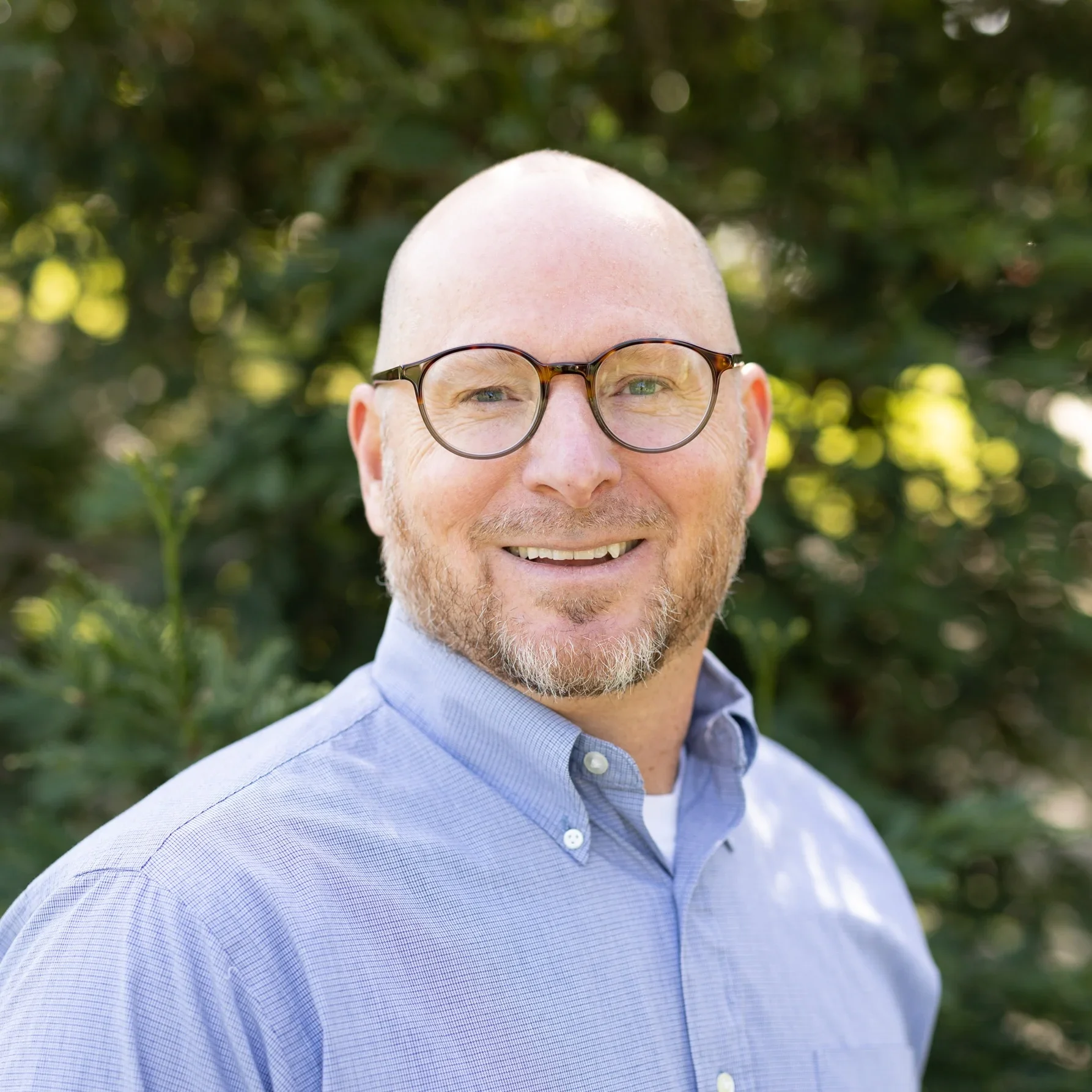 A smiling man with glasses and a beard, wearing a light blue button-down shirt, standing outdoors with greenery in the background.