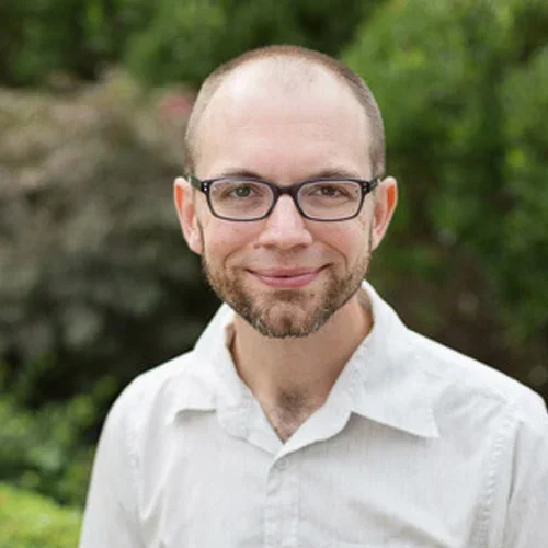 A man with glasses, a beard, and short hair smiling outdoors with green trees in the background.
