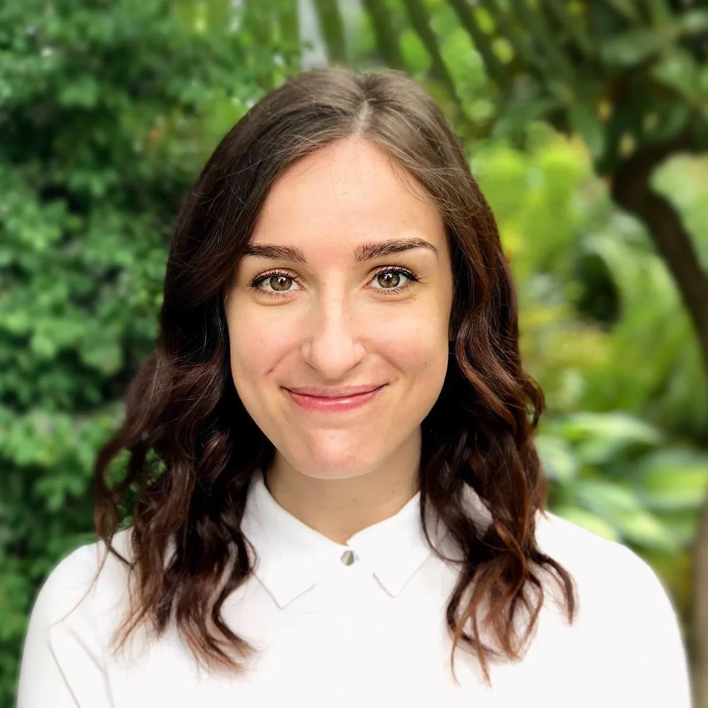 Young woman with shoulder-length wavy brown hair outdoors, smiling, wearing a white collared shirt, with green foliage in the background.