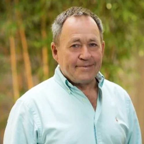 A middle-aged man with short gray hair wearing a light blue shirt outdoors with green foliage in the background.