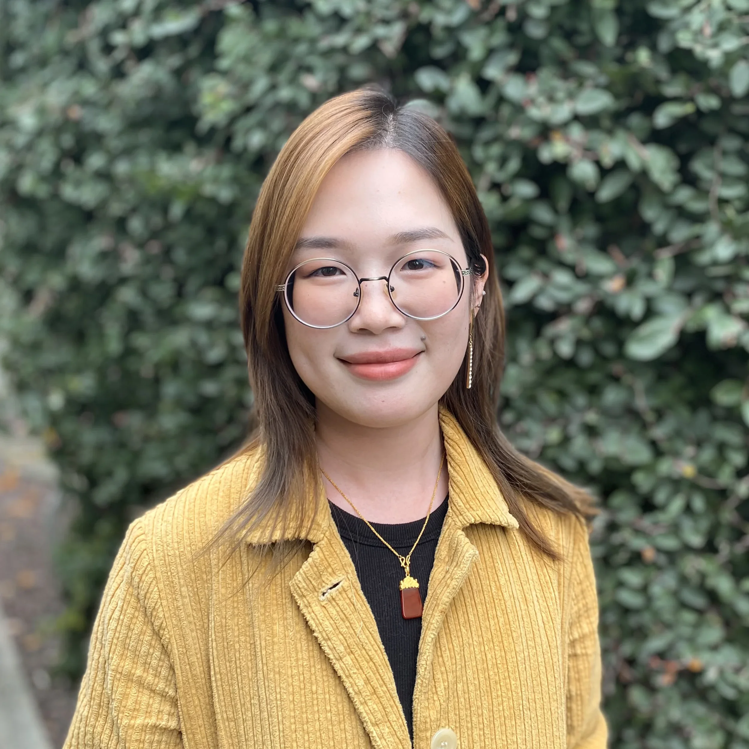 A young woman with shoulder-length brown hair, wearing glasses, a mustard yellow jacket, and layered gold jewelry, standing outdoors in front of green foliage and smiling at the camera.