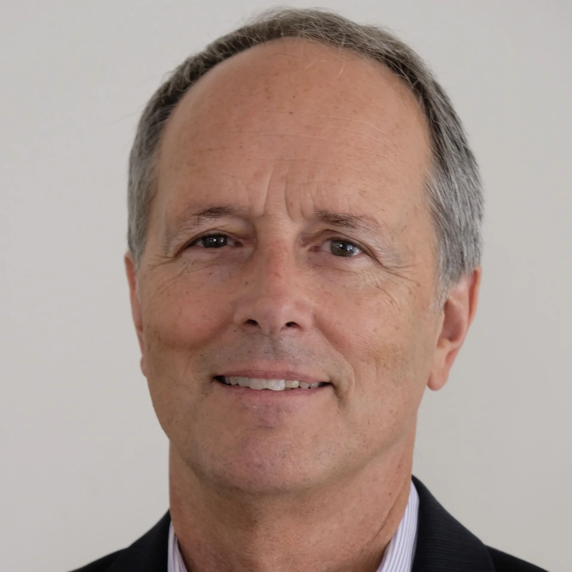 Close-up portrait of smiling middle-aged man with short gray hair, wearing a dark suit and light-colored shirt, against a plain light background.