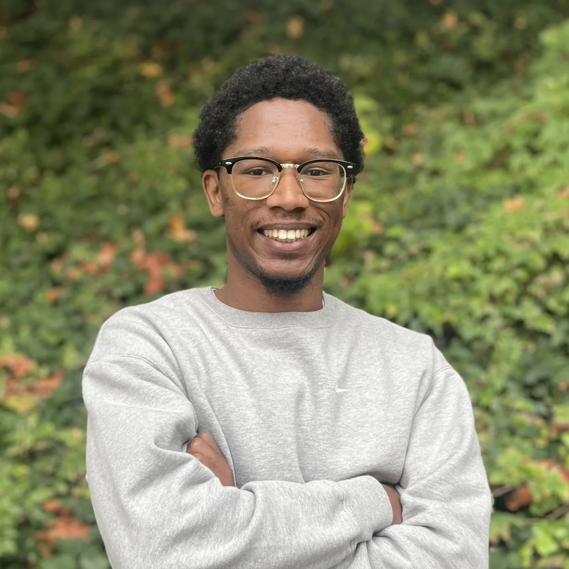 A young man with glasses smiling outdoors against a blurred background of green and autumn leaves.