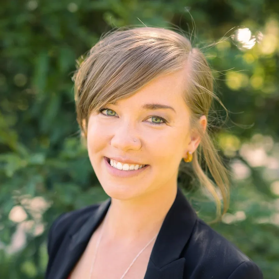 Smiling woman with short brown hair and green eyes outdoors with blurred green foliage in the background.