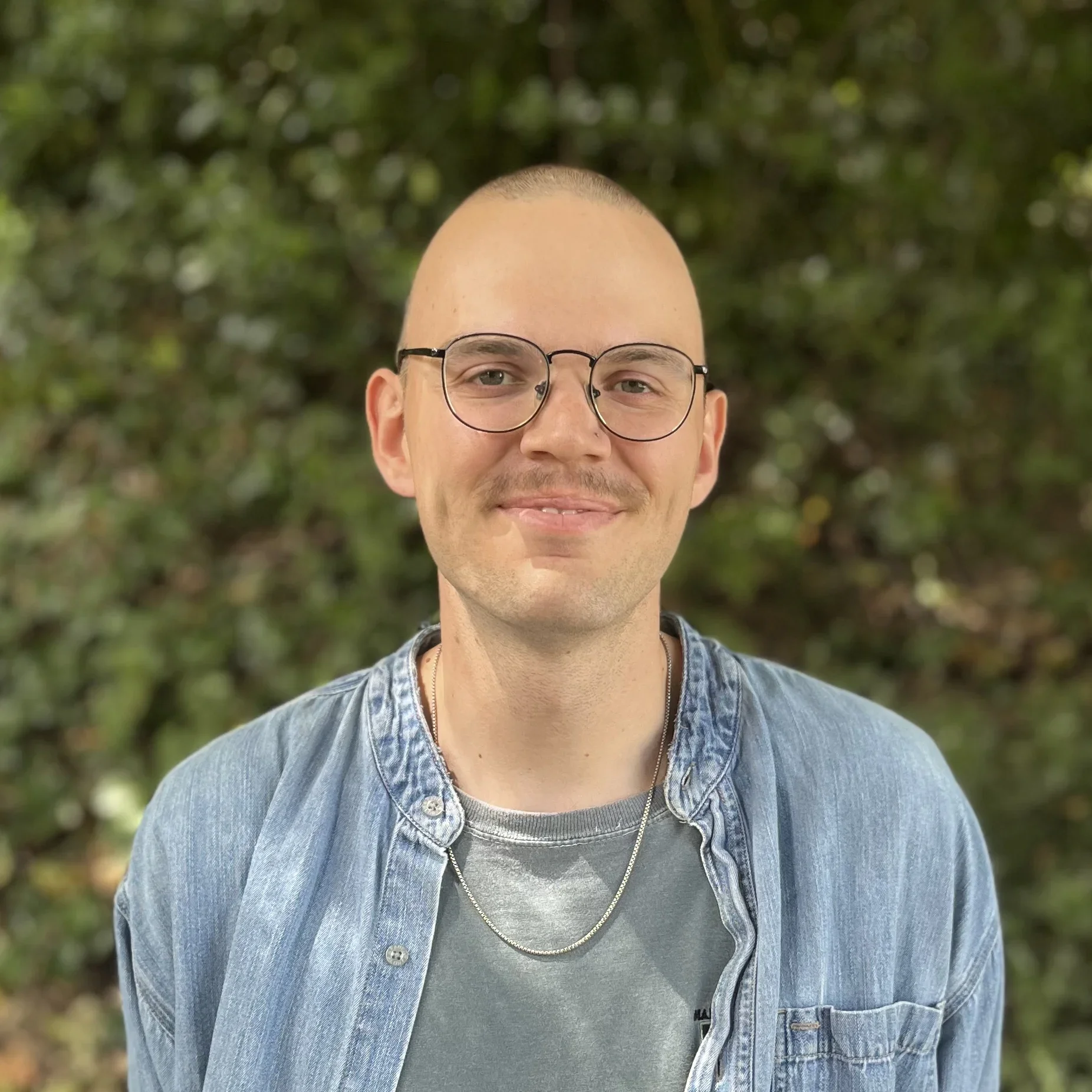 A young man with glasses, a shaved head, wearing a denim jacket and a chain necklace, standing outdoors with green foliage in the background.