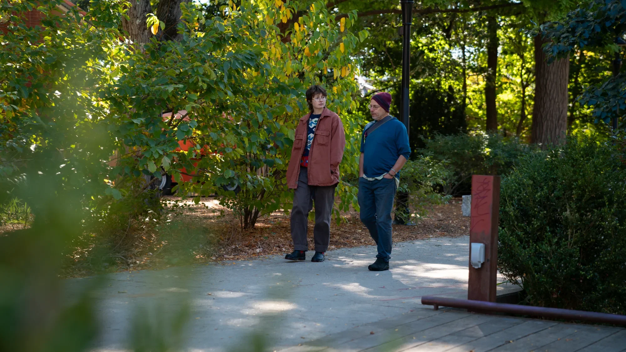 Two people walking and talking on a shaded park path surrounded by trees and bushes.