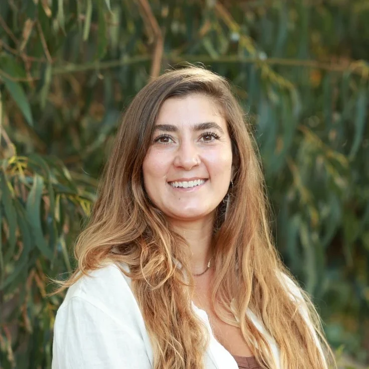A smiling woman with long, wavy brown hair standing outdoors in front of green foliage.