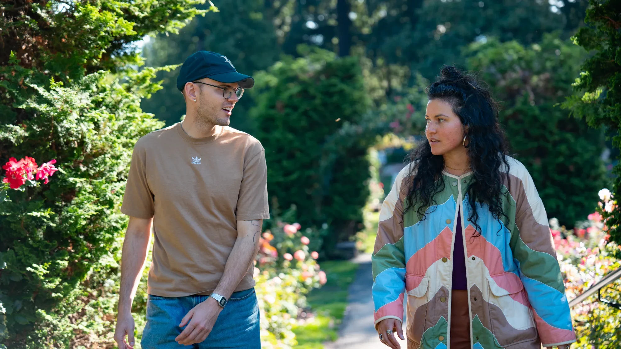 A man and a woman walking and talking in a lush garden with colorful flowers and trees.
