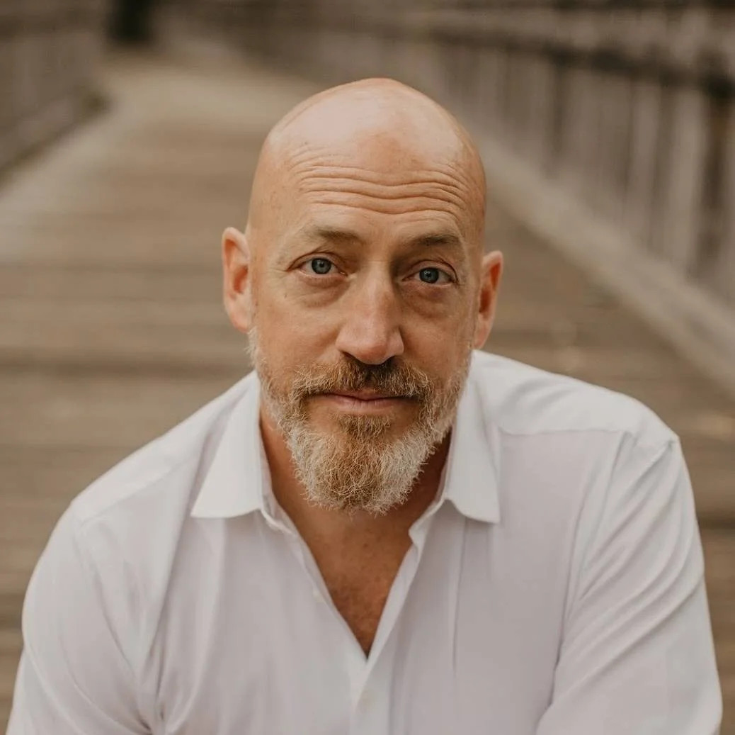 A man with a bald head, blue eyes, and a beard, wearing a light blue shirt and a tan jacket, smiling outdoors with greenery in the background.