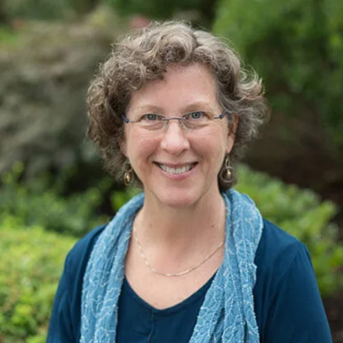 A smiling woman with curly gray hair, glasses, wearing earrings, a blue top, and a blue scarf, standing outdoors with green foliage in the background.