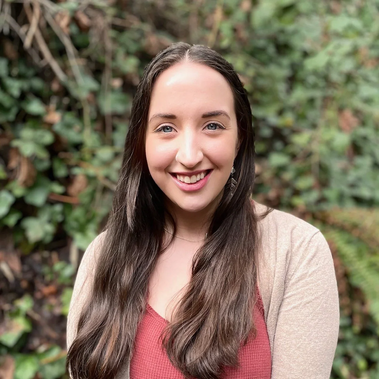 A woman with long dark hair smiling outdoors with green foliage in the background.