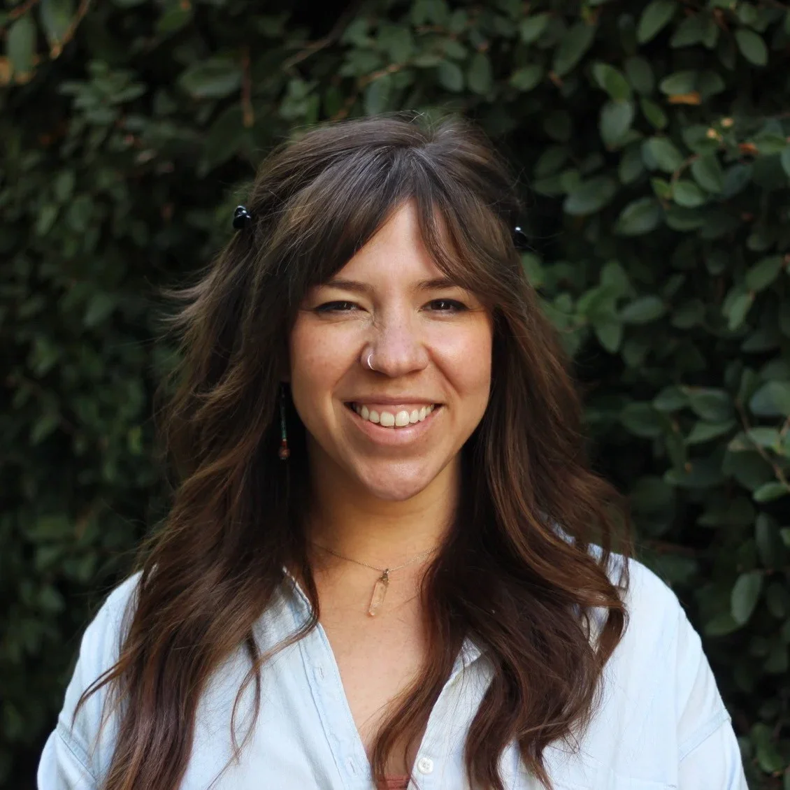 A woman with long brown wavy hair, wearing a light blue shirt, standing outdoors with a green leafy background, smiling at the camera.