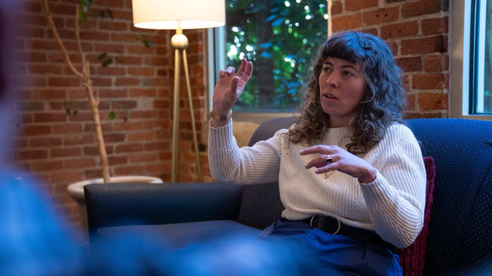 Woman with curly hair sitting on a sofa in a room with brick walls and a window, gesturing with her hand as she talks.