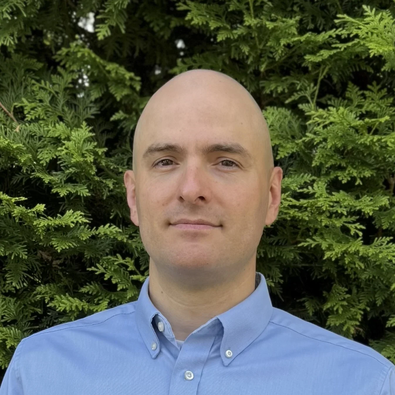 Portrait of a bald man with light skin, wearing a light blue collared shirt, standing outdoors in front of green leafy bushes.