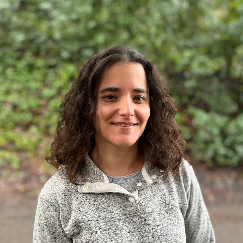 A woman with wavy brown hair smiling outdoors with green foliage in the background.