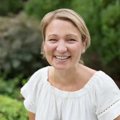 A woman with short blonde hair smiling outdoors with green foliage in the background.
