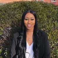 A woman with long black hair, wearing a black blazer and white top, standing outdoors in front of greenery.