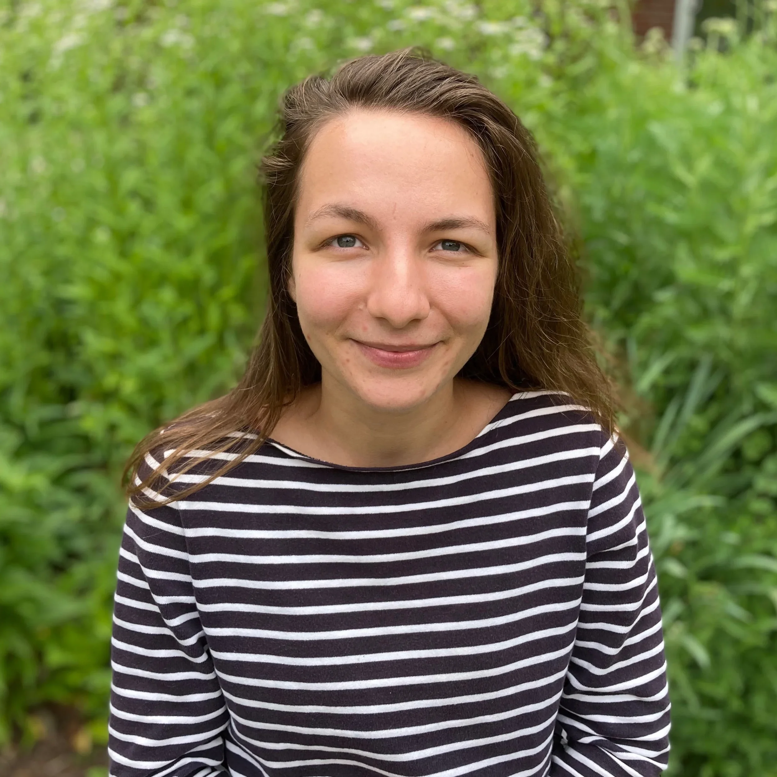 A young woman with long brown hair, light skin, and blue eyes, smiling in front of green foliage, wearing a black and white striped shirt.
