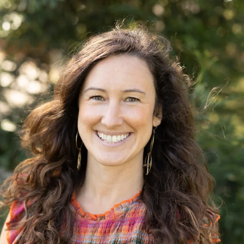 A woman with wavy brown hair smiling outdoors with greenery in the background.