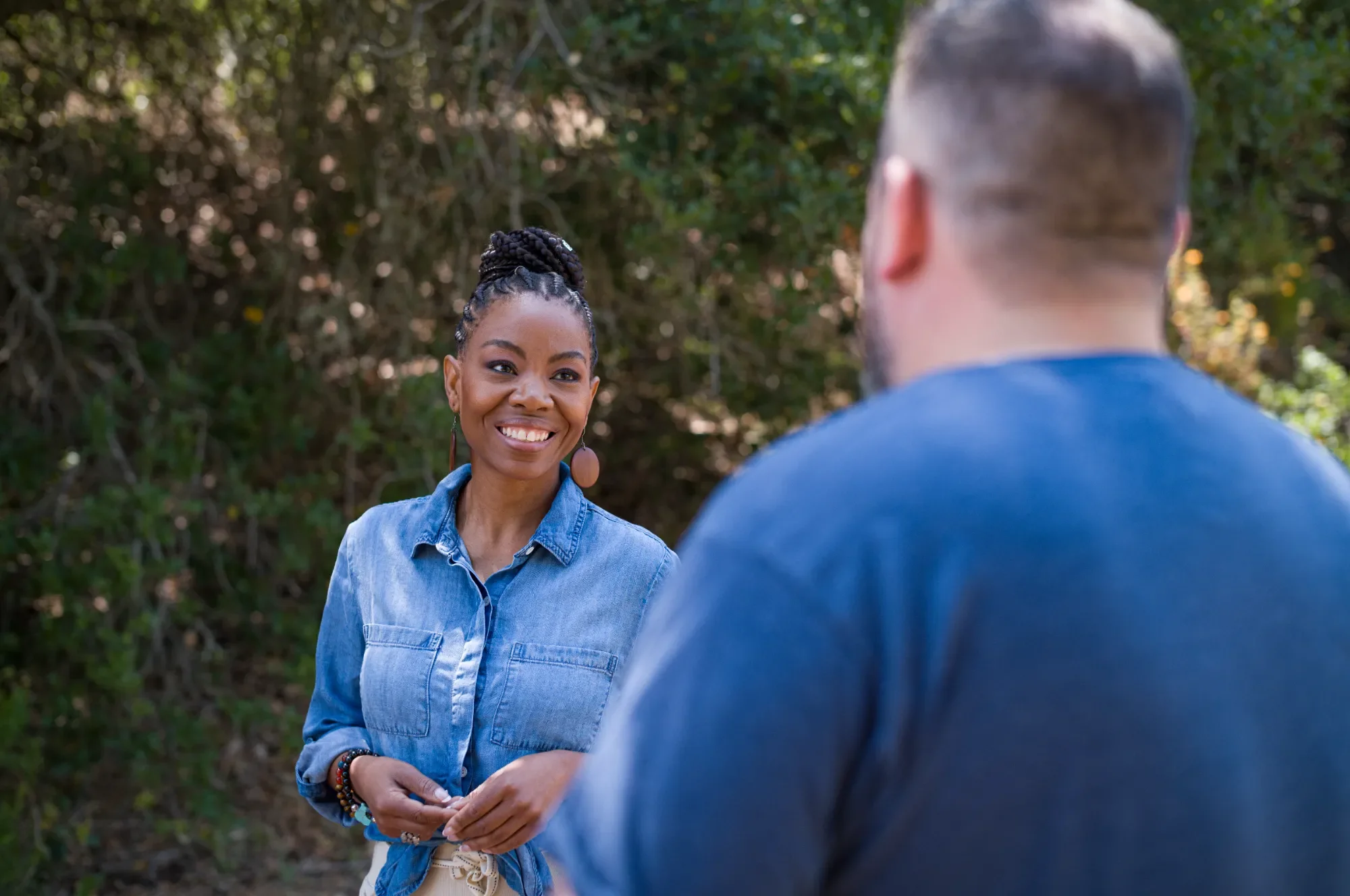 A woman smiling and talking to a man outdoors with green trees in the background.