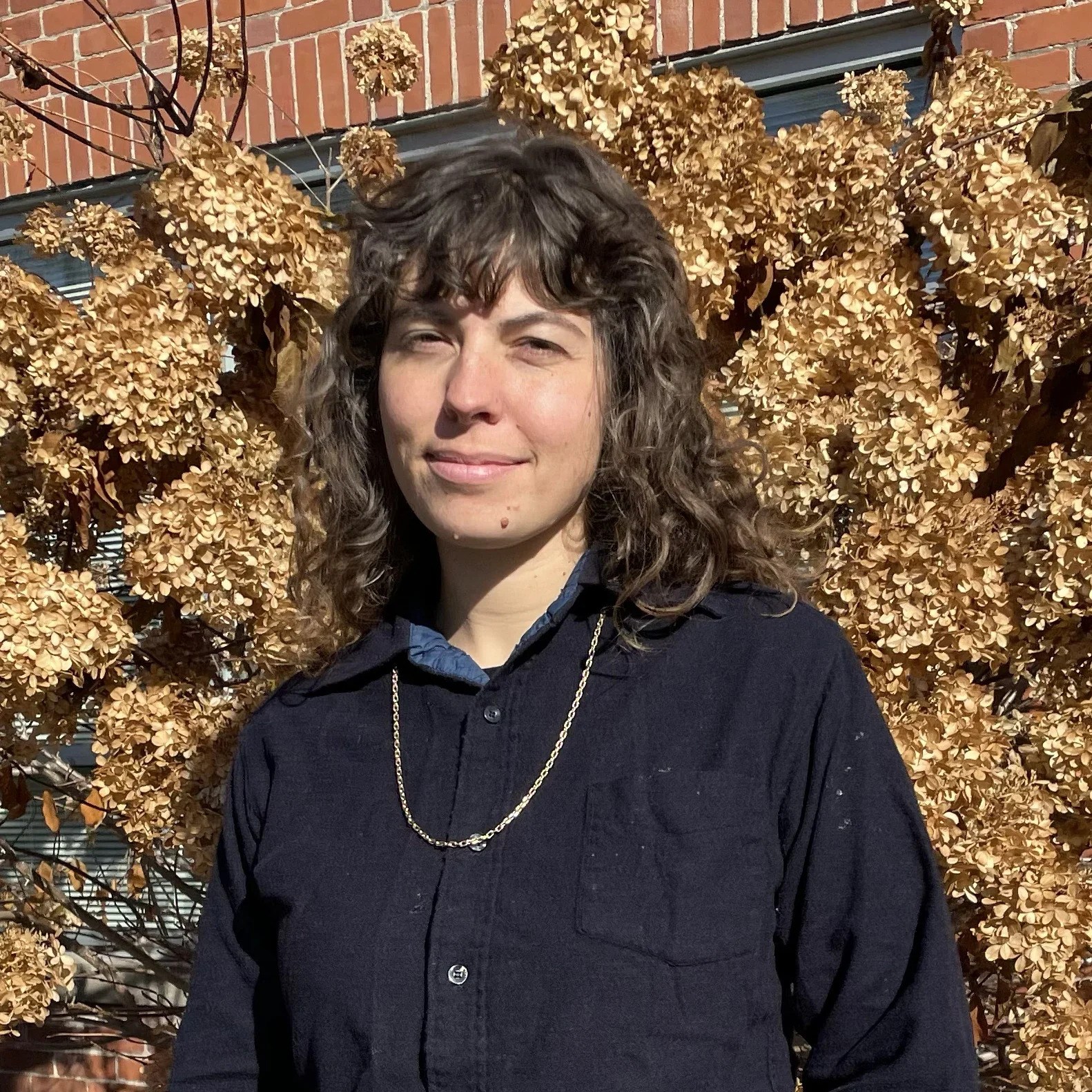 A woman with curly brown hair standing outdoors in front of a bush with dried, brown hydrangea flowers. She is wearing a dark jacket over a blue shirt and a gold chain necklace. She is squinting slightly in the sunlight.